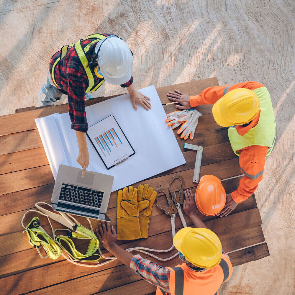 Overhead shot of engineers around a table with tools and gloves, papers and a laptop.